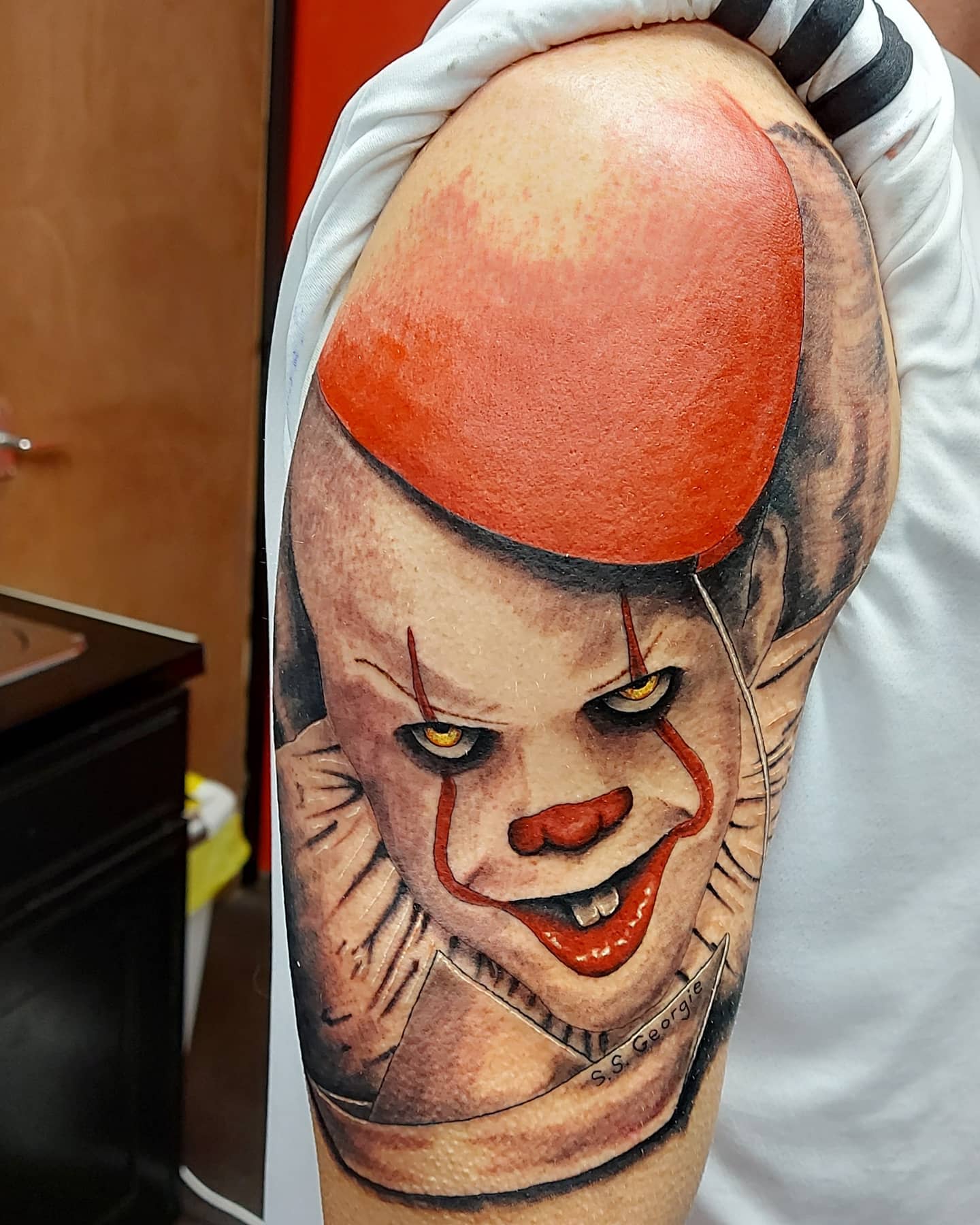 Realistic Pennywise tattoo on upper arm, with a large red balloon and Georgie's paper boat